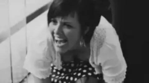 Family A black and white image of a woman kneeling down and laughing. She has short, dark hair, large hoop earrings and is wearing a white top over a dark spotty dress.