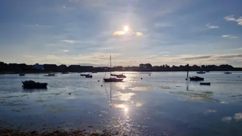 Emsworth Harbour on a sunny day with several boats moored in the water The sun is in the sky overhead and reflects brightly in the water. Behind the harbour you can see buildings from the town.