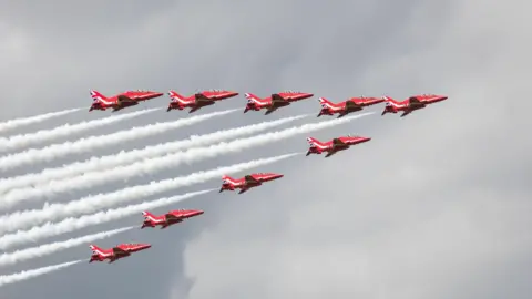 Hang Ross Nine Red Arrow jets in a triangular formation, leaving smoke trails in their wake. The red planes have Union Jack livery on their tails.