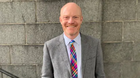 A man in his 40s stood in front of a brick wall wearing a white shirt, Multi-coloured tie and grey suit jacket.