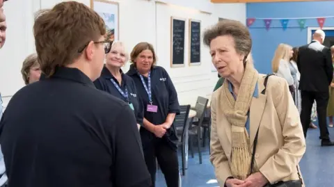 An older woman with pinned back brown hair, wearing a pale brown jacket and scarf with a handbag over her shoulder, stands in a room with a blue floor and white walls, talking to a row of people all wearing dark blue tops with lanyards round their necks. There are some people in the background and some colourful bunting on a blue wall.