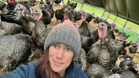 Postance Poultry Kate Postance of Postance Poultry takes a selfie in the the purpose-built tunnel housing her turkeys.  The turkeys can be seen behind her.  Kate has long brown hair and is wearing a grey, woollen hat and a blue fleece.