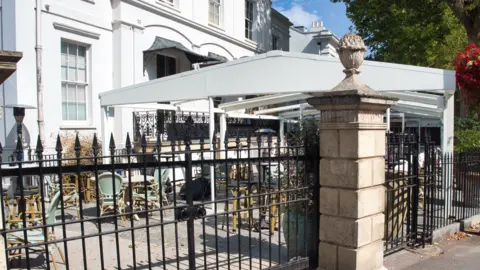 Cheltenham Civic Society A large pergola line the front of a historic building. They are square with shallow pointed roofs, in white metal. 