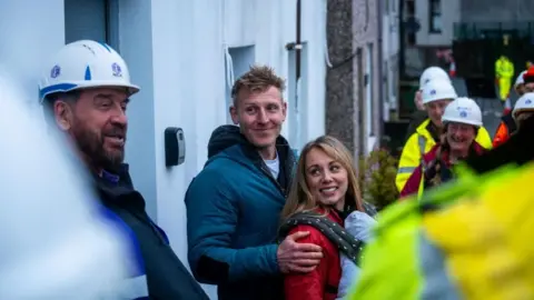 South Shore People in hard hats, stood outside a house. In the centre of their gathering are a man and a woman. The man, with short blonde hair, is wearing a blue hooded jacket. he is smiling and has his hands on the shoulders of a blonde woman in front of him, who wears a red jumper and a polka-dot baby carrier on her front, with the outline of an infant visible inside it. She is also smiling and they are both looking at one man, with a beard, in a white hard hat.