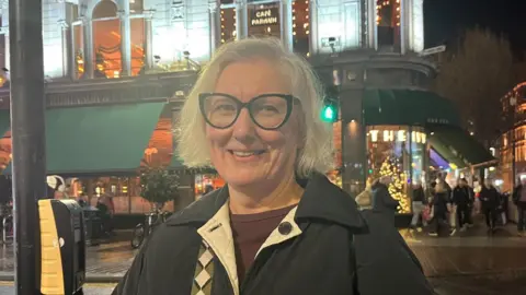 Ms Adair has shoulder length blonde hair and black glasses. She is wearing a navy coat and burgundy top.