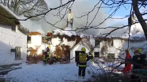 The picture shows a group of firefighters tackling a major blaze at a traditional thatched-roof building. Thick smoke is billowing from the roof, which appears to be severely damaged and partially collapsed. Several firefighters in bright yellow helmets and high-visibility jackets are using ladders and hoses to control the fire, while foam or water covers the ground in front of the building. The words “Ring of Bells” are visible on the front wall.