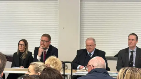 Community meeting attended by Jonathan Brash, Hartlepool Councillors Melanie Morley and Moss Boddy and Stockton North MP Chris McDonald. All four of these are pictured wearing dark black/grey suits. They are sat at a long table in front of an audience. A few members of the audience are in the image, but only the backs of their heads are visible.