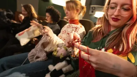 Women sit in a cinema and knit and crochet. 