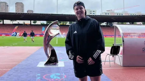 Abby Morton Abby Morton standing next to the UEFA Women's euros silver trophy. She has a black Adidas jacket on and black shorts. Morton is smiling.