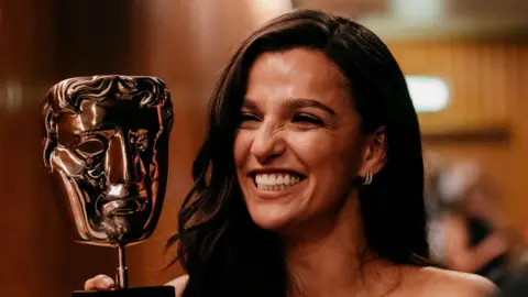 A woman with long dark hair, smiling as she looks at a gold Bafta TV award next to her head