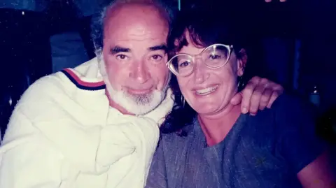 Eli Getreu is seen here with his wife Barbro Gentele. They are sitting close together and Mr Getreu has his arm around Ms Gentele. He is wearing a white top with a white jumper around his shoulders. She is wearing glasses and a blue dress and a watch.