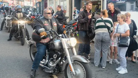 A picture from Grantham Bike Night 2025. A large silver motorbike is at the centre of the picture. It is being ridden by a man, with the headlamp on. Two other bikes follow it in procession. 