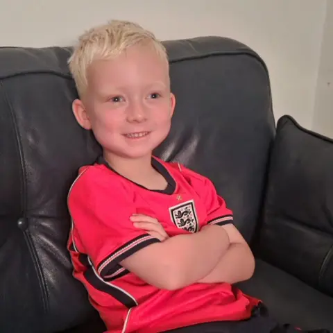 Nicola Finlay is wearing a red England shirt with dark blue trim and the team badge on it. He is sitting on a black leather sofa and smiling at the television. 