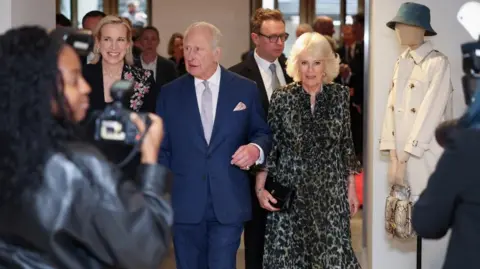 Reuters King Charles and Queen Camilla walk together into a reception as people mill about in the background and with media in the foreground. He is in a blue suit and pale shirt and tie, She is in a green and black patterned dress.
