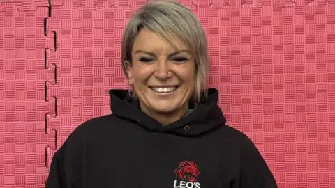 Leo's Gym Head and shoulders image of coach Carrie Bradburn smiling in front of a wall of pink rubber gym floor matting. She is wearing a black hoodie with a red lion logo and has short blonde hair, which is longer on one side
