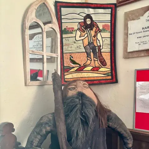 Rory Waterman A papier-mache depiction of a man with a long brown beard and brown hair is perched on a church seat holding a club on his shoulder. Above it a rectangular felt drawing is hanging on the wall next to a church-window shaped mirror.