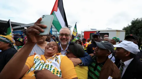 Reuters A smiling Ebrahim Rasool stands in a crowd of supporters at Cape Town airport. He is being hugged by a woman, while another suppoter takes a selfie with him.