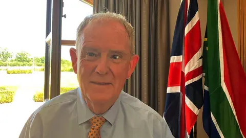Head and shoulders of Professor Stephen Grigg standing beside the Union flag and South Africa's flag by the window in his house. He has short white hair and is wearing a pale blue shirt and yellow patterned tie