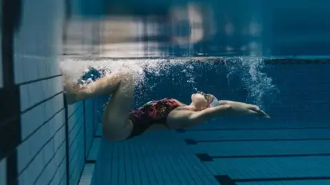 Getty Images A female swimmer in a black, pink and purple costume is underwater and about to push off from the side of a swimming pool to do backstroke, with her arms outstretched.