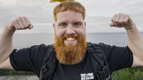 A man with a big ginger beard smiling and flexing his arm muscles. 