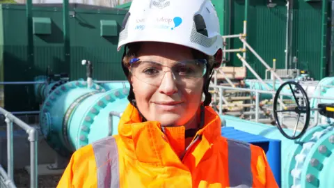John Fairhall/BBC A woman wearing an orange hi-vis jacket and a white helmet. She is smiling at the camera and wearing safety goggles 