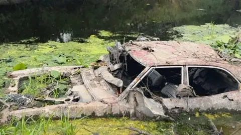 A rusted, rotten car overgrown with weeds and surrounded by algae sits in the water of the River Lagan. The wheels are submerged beneath the water but the body of the car is entirely visible. The windows are down. It's a dusty orange-pink colour.