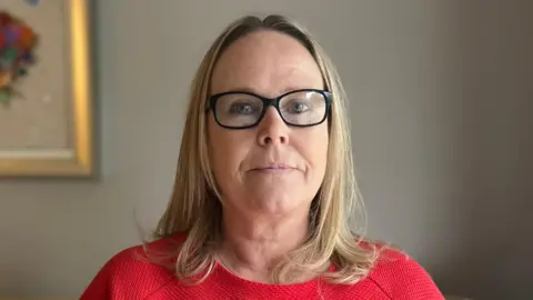 A middle-aged woman is looking down the lens of the camera, she has shoulder-length blonde hair and is wearing black-rimmed glasses and a red top. She is inside, possibly in the living room of a house. In the back ground is a grey wall on which we can see the corner of a piece of framed artwork. 