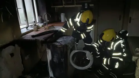 Jersey Fire Two firefighters in protective clothing and wearing yellow helmets inspect a burnt tumble dryer following a blaze which has caused significant fire damage to the room it is in.