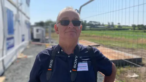 A man with short white hair wearing sunglasses and a dark blue short-sleeved shirt. He is standing outside with metal fencing in the background.