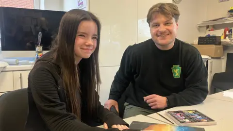 BBC / ELIZABETH BAINES A 14-year-old girl with long, brown hair smiles at a table covered in artwork. A man in his 30s smiles beside her. He wears a black sweatshirt bearing the green and gold logo of The Hunslet Club.