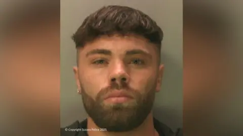 Sussex Police A police mugshot of a young man. He has a beard and brown hair. 