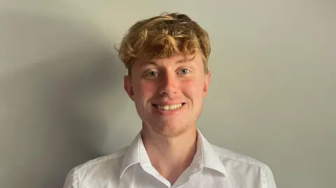 Gloucestershire Community rail Partnership A photo of a white man with blond hair and wearing a white shirt. He is stood in front of a grey wall