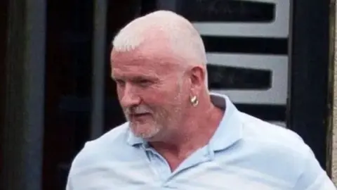 Pacemaker Malcolm McKeown has cropped grey hair and is wearing an ear ring in his left ear. He is wearing a light blue open-necked shirt