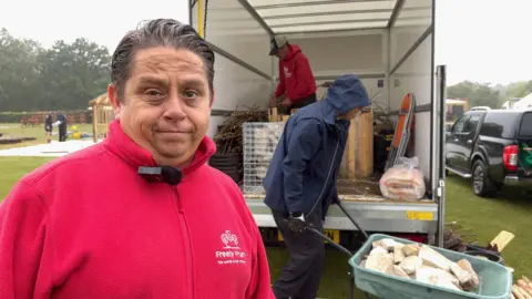 A man in a red fleece grimaces at the pouring rain while a colleague, hood up against the weather, pushes a wheelbarrow full of decorative stone.