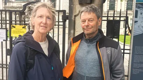 A grey haired woman and man wearing outdoor wear and backpacks stand in front of the gates of Belfast City Hall