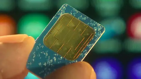 A close up image of a small gold coloured mobile phone SIM card with a smart phone out of focus behind it. 