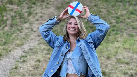 A young woman with blonde hair holds a wheel of Double Gloucester on her head as she stands at the bottom of Cooper's Hill. She is wearing a blue denim jacket and is smiling.