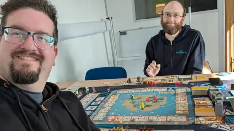Rikh Hart Rikh on the left is taking a selfie with Stuart. They are sitting at a table with a complicated board game at play in front of them