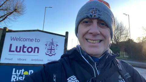 A man is smiling in front of a Welcome to Luton sign and wearing a Luton Town branded hat.