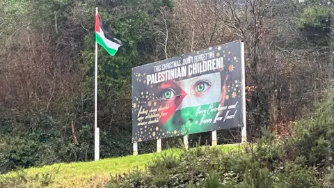 The picture shows a billboard up on a grassy hill saying "This Christmas don't forget Palestinian children" with a Palestinian flag beside it