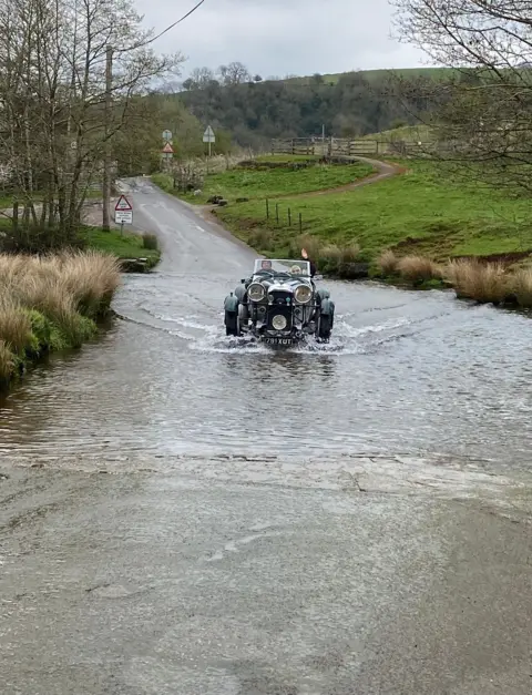 Megan Waugh An old motor car passes through a flooded road