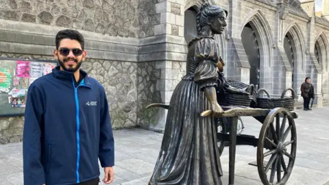 A man with sunglasses and a navy coat stands beside the famous statue of Molly Malone.