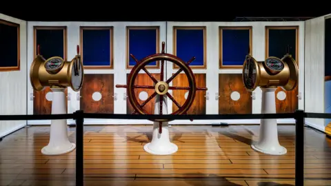 A wooden ship's wheel and compasses on display on a fake wooden floor with a fake ship display behind it.