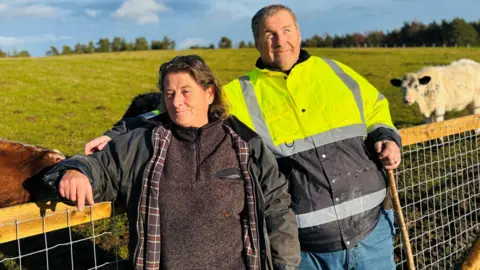 Ness and Mike Duxbury smiling at camera, she is wearing farming-style clothing, he is wearing a bright yellow safety jacket, there are cows in a field in the behind them.