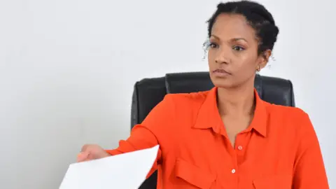 A woman hands over a document in an office. She is wearing an orange shirt and has black, short hair. 