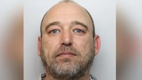 A mug-shot of Michael Sergio Smith, looking straight at the camera, he has blue eyes, short hair, and a beard. 