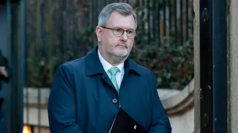 PA Media Sir Jeffrey Donaldson, a man with grey hair and dark glasses, wearing a blue jacket, white shirt and green tie, holding a black and red book.