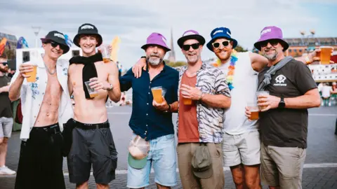Nadine Ballantyne A group of men in a huddle looking at the camera. They are all wearing bucket hats and holding plastic cups full of beer. They are all smiling at the camera