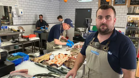 A man wearing an apron in a fishmongers. Behind the man is a table covered in fish on ice and workers in aprons, preparing fish.