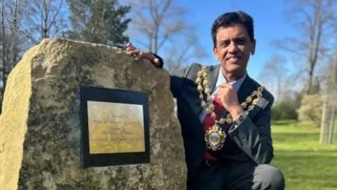 The Mayor of Oldham Dr Zahid Chauhan rests on his haunches next to the memorial boulder. He is wearing his mayoral chain. The boulder has in inscription on it that cannot be made out in the photograph. 